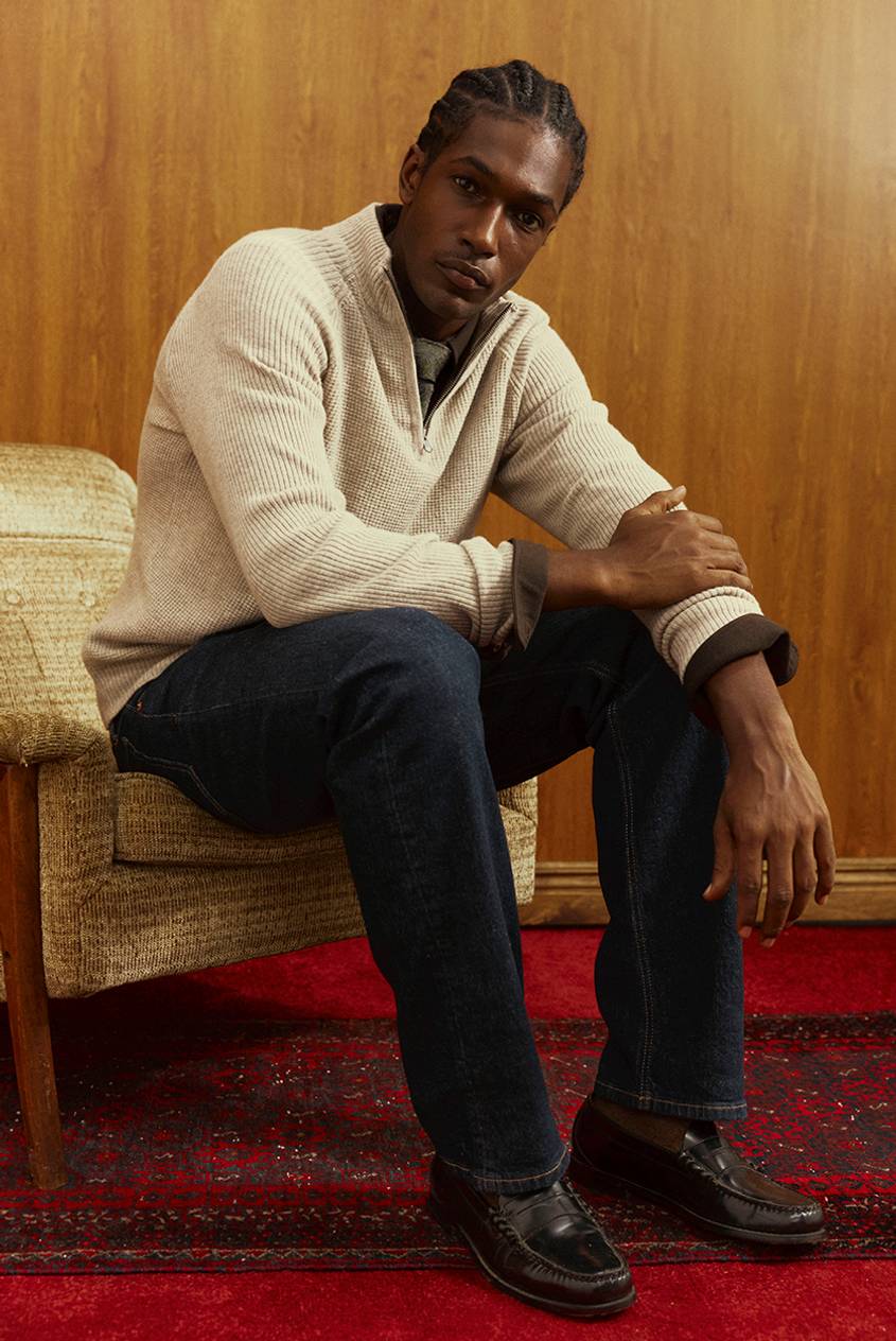 A man sits in a vintage armchair wearing a cream ribbed half-zip sweater over a collared shirt, dark denim, and brown leather loafers, with wood paneling and a red patterned rug behind him.