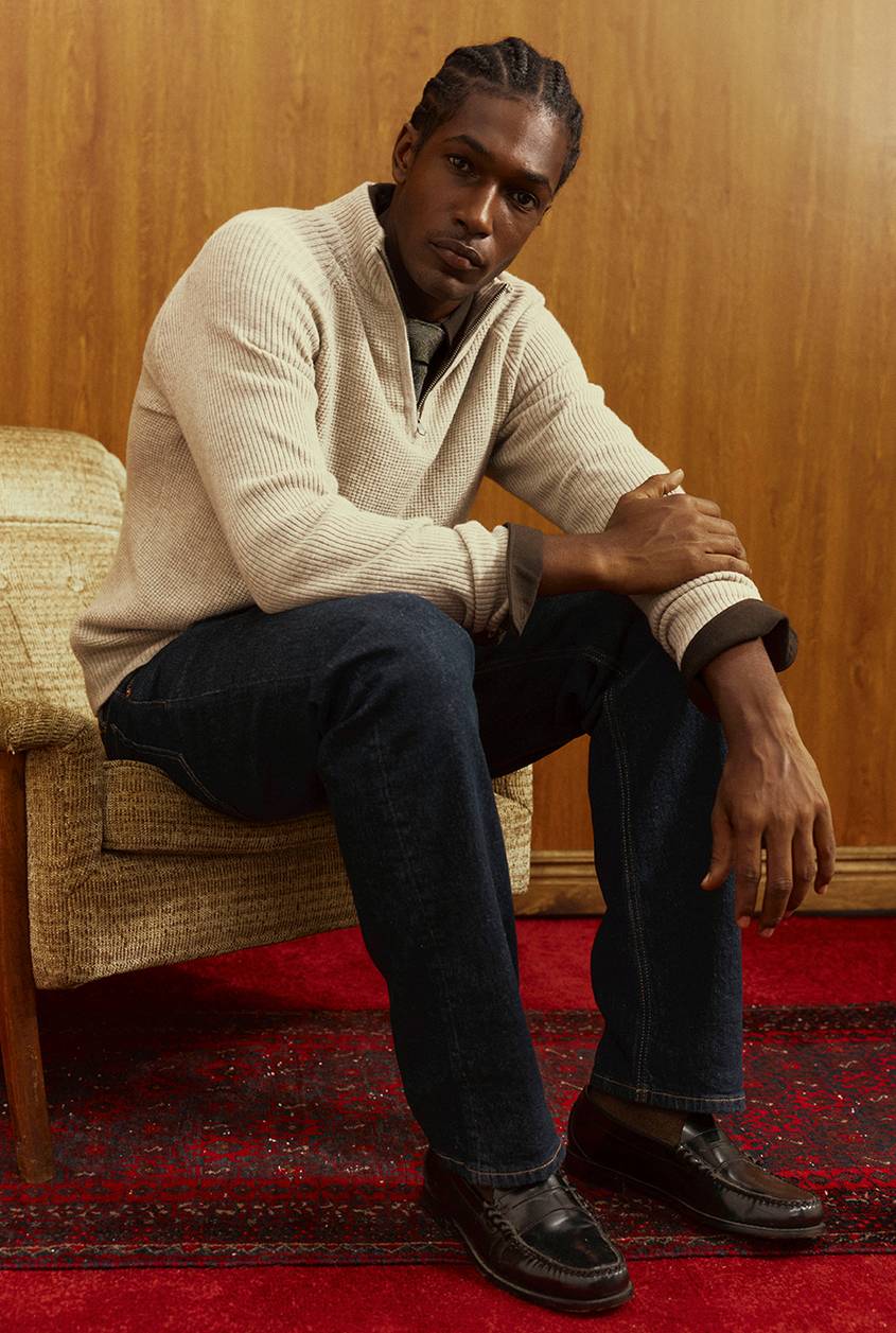 A man sits in a vintage armchair wearing a cream ribbed half-zip sweater over a collared shirt, dark denim, and brown leather loafers, with wood paneling and a red patterned rug behind him.