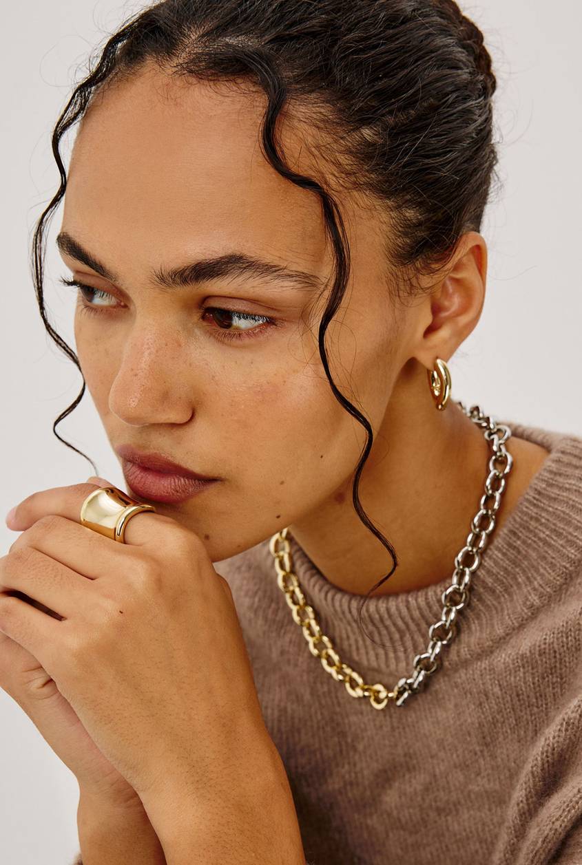 Close-up of a model wearing layered gold and silver chain necklaces and gold hoop earrings with a taupe knit sweater.