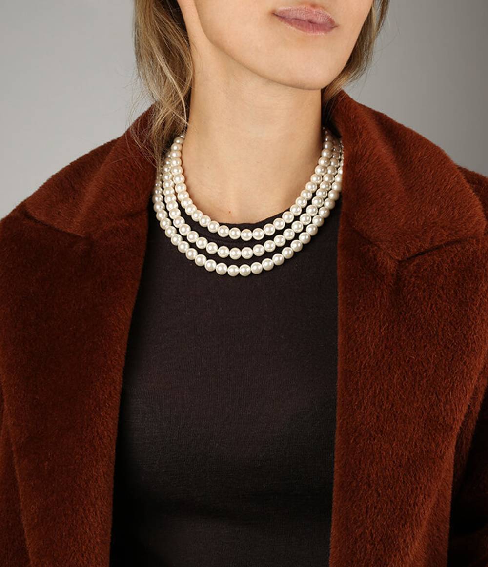 A girl with a brown coat on is wearing a three strand white shell pearl necklace.