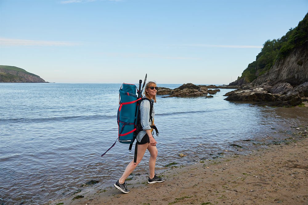 woman walking on beach carrying paddle board bag