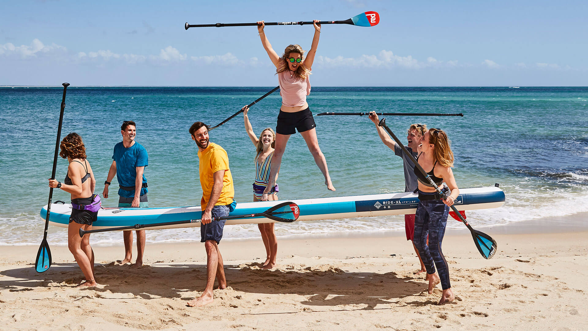group of paddlers carrying multi-person paddle board into the water with a woman stood on top