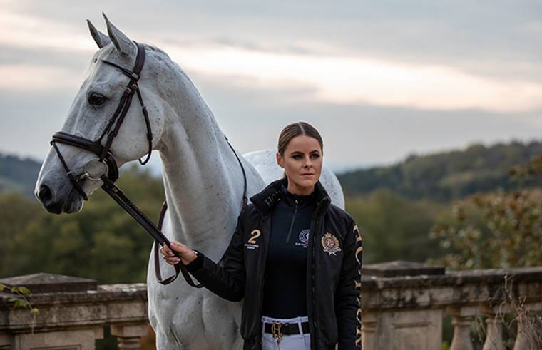 Jade Holland Cooper with white horse holding bridle dressed in HC Team Jacket and Black Base Layer