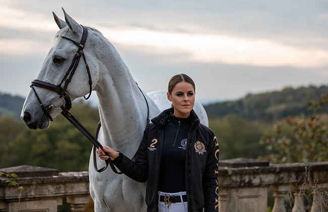 Jade Holland Cooper with white horse holding bridle dressed in HC Team Jacket and Black Base Layer