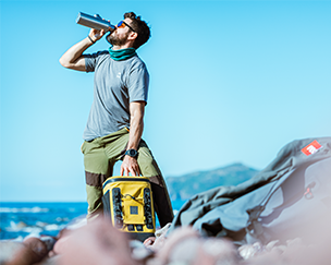 Man drinking from Red Original insulated water bottle whilst holding a cool bag backpack in mustard