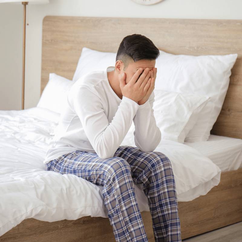 man sitting on the edge of a white bed wearing pyjamas and holding his head in his arms because of sleep deprivation