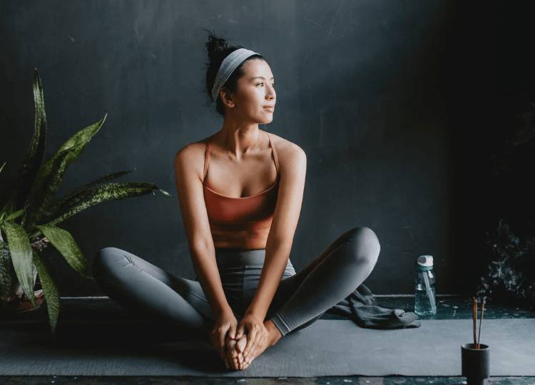 women performing a seated yoga post whilst sitting on a yoga mat and looking into the distance as if contemplating and relaxing in that moment at the same time