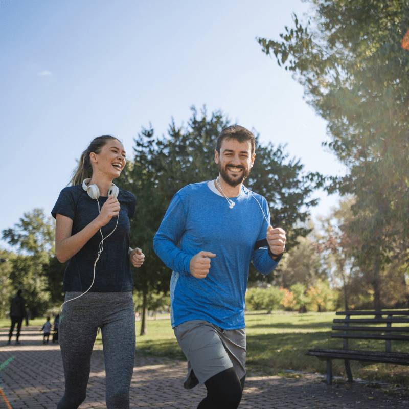 woman in black sports clothing and man in blue sports clothing running together outside in a park in the sunshine laughing and smiling