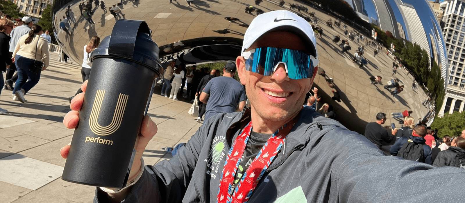 Runner Craig Sharp posing with a U Perform Shaker Bottle after finishing the Chicago Marathon, wearing his finishers medal and blue tinted sports sunglasses