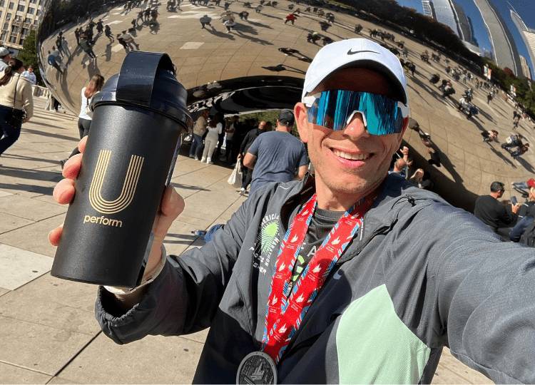 Runner Craig Sharp posing with a U Perform Shaker Bottle after finishing the Chicago Marathon, wearing his finishers medal and blue tinted sports sunglasses