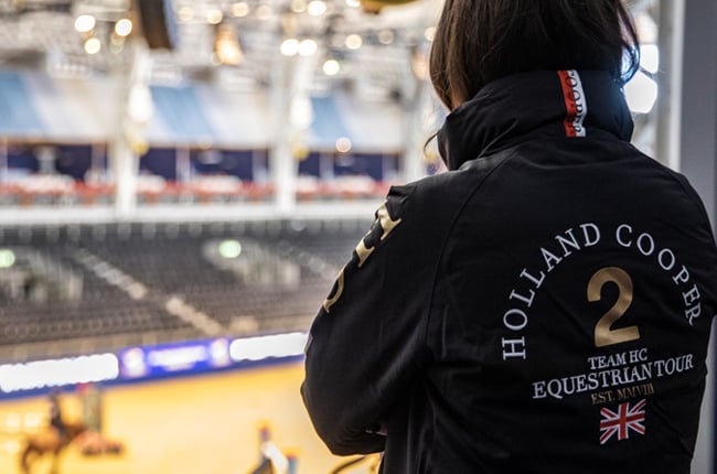 Woman stood looking away from the camera wearing a navy blue team jacket with text on the back saying 2 Holland Cooper with a union jack