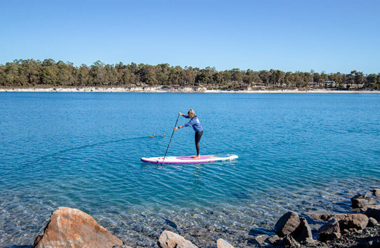 woman paddle boarding on a blue lake with large trees in the background