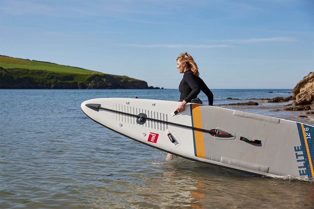 Woman carrying Red Original racing SUP into water on the beach