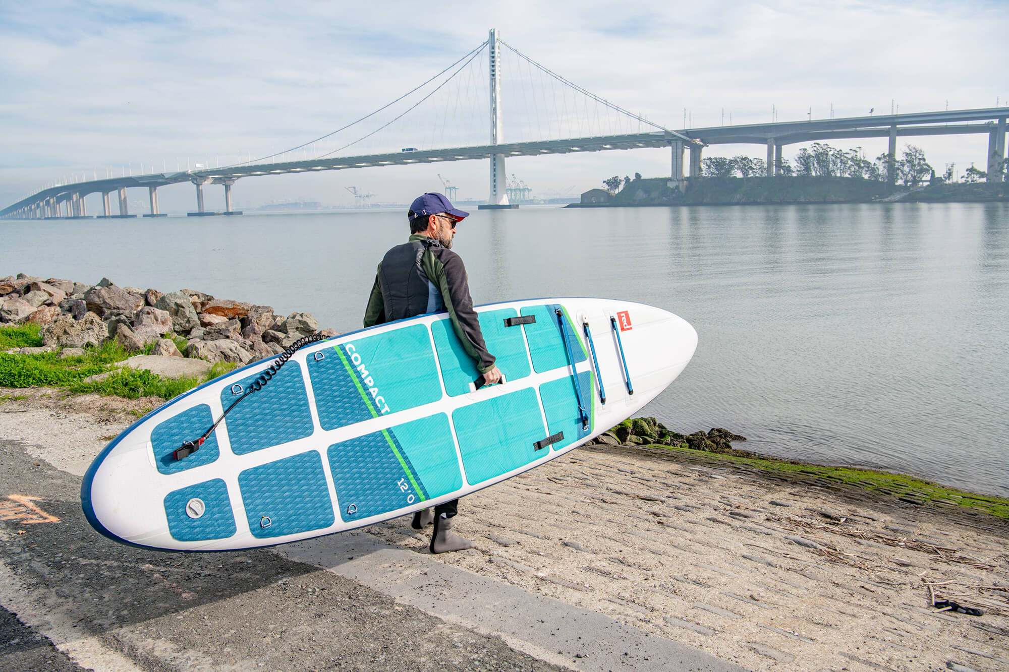 Man Holding An RPC 12 Compact SUP On The Shore In front Of The San Mateo–Hayward Bridge