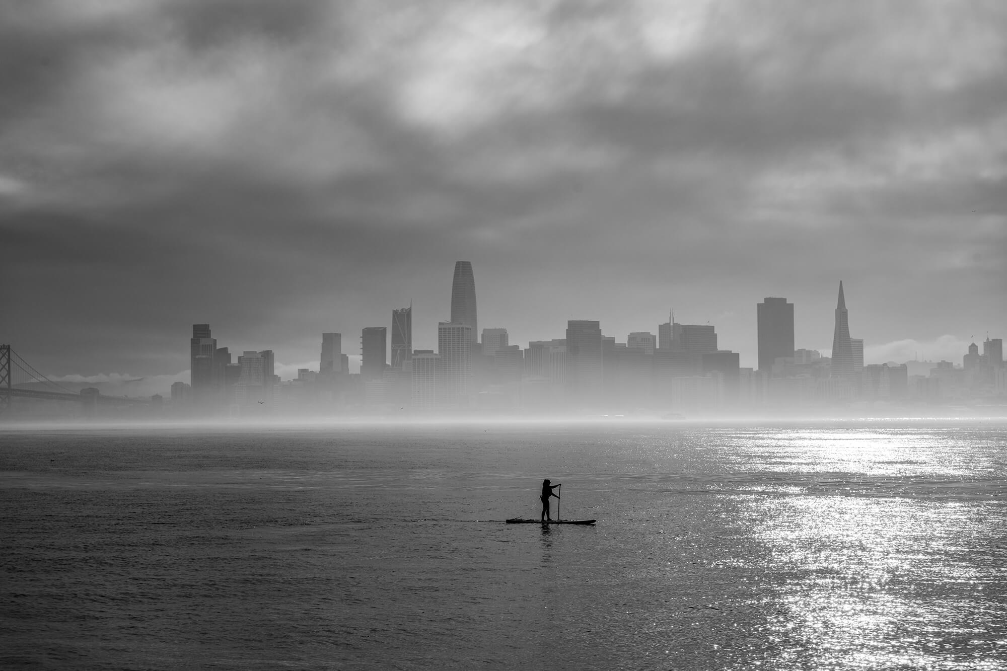 Woman Paddle Boarding On A Foggy Day With The Silhouette Of The San Francisco Cityscape Behind Her