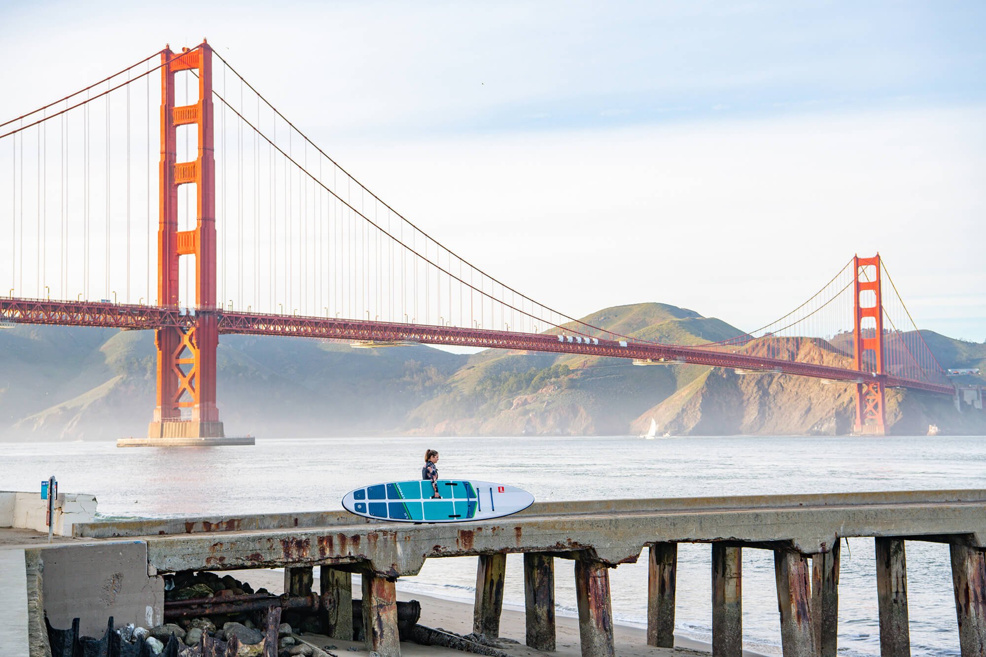 Woman Carrying A Red Paddle SUP Along A Jetty In Front Of The Golden Gate Bridge