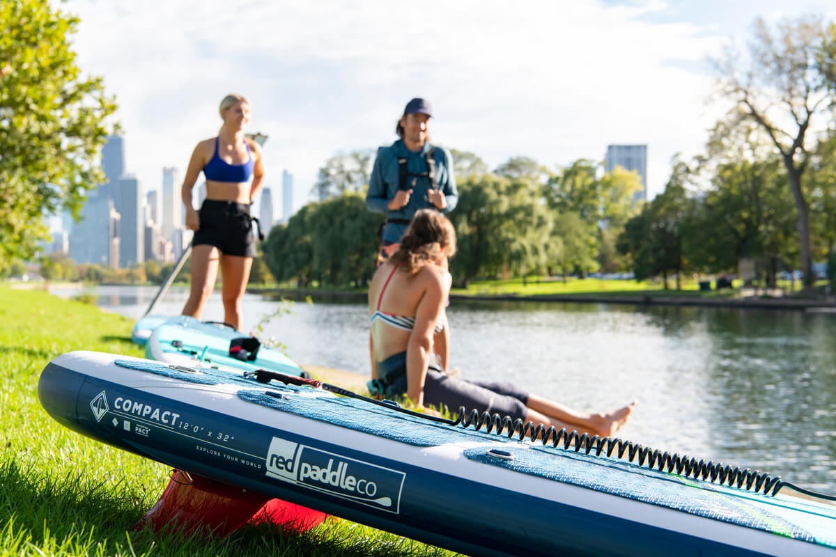 People sat on a grassy bank by a river next to a Red Paddle Co Compact Paddle Board