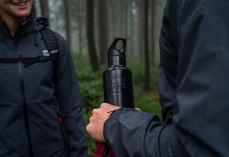 Close up of person holding a reusable water bottle in a forest whilst talking to someone else
