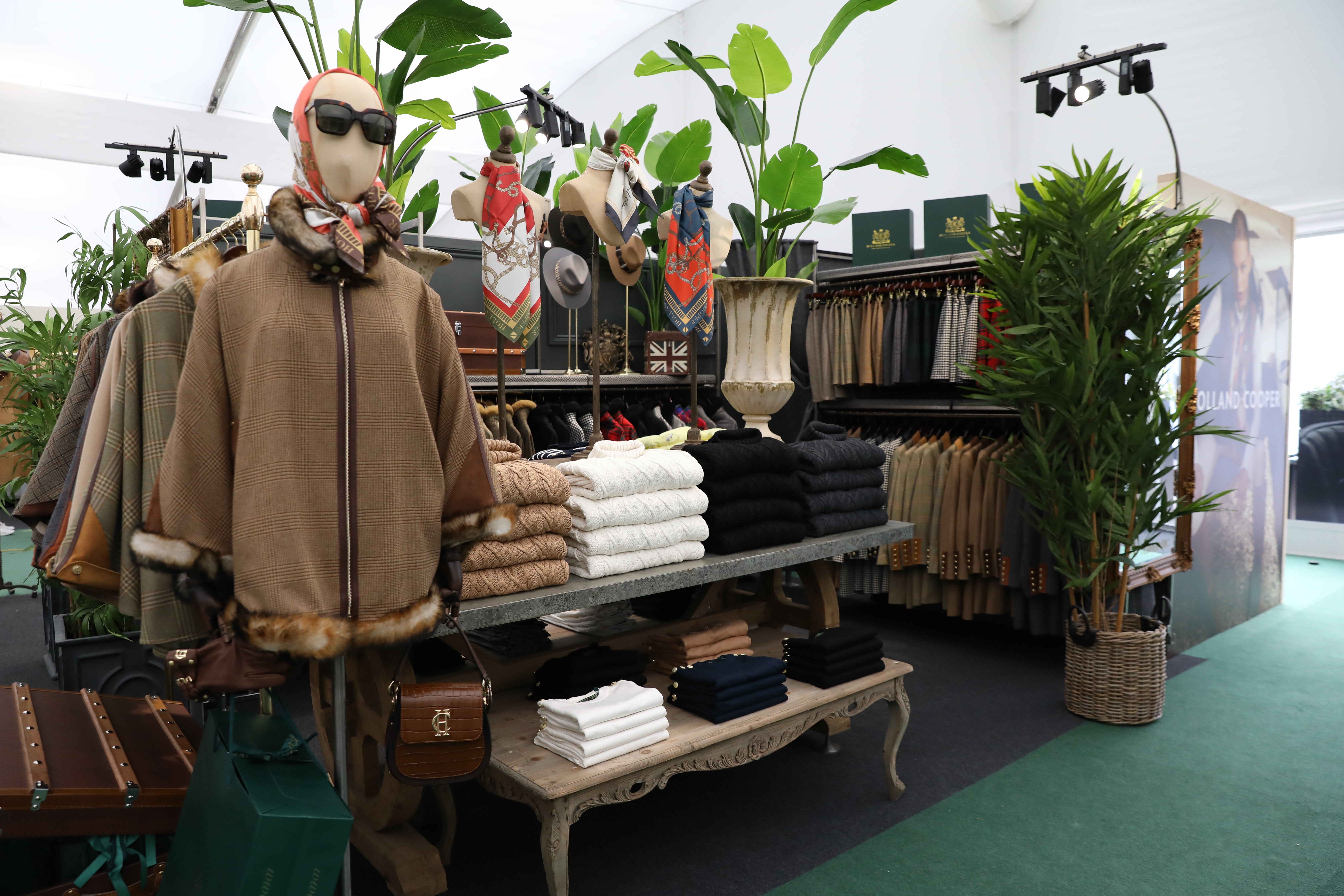 Store layout with clothing folded on tables and mannequin wearing brown tweed cape, black sunglasses and red silk scarf wound the head