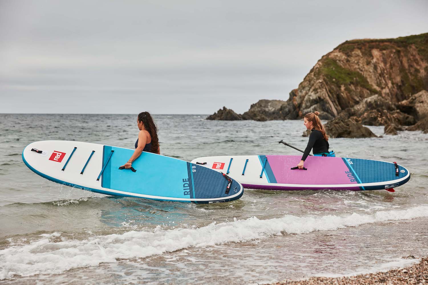 two women carrying paddle boards wading into the sea