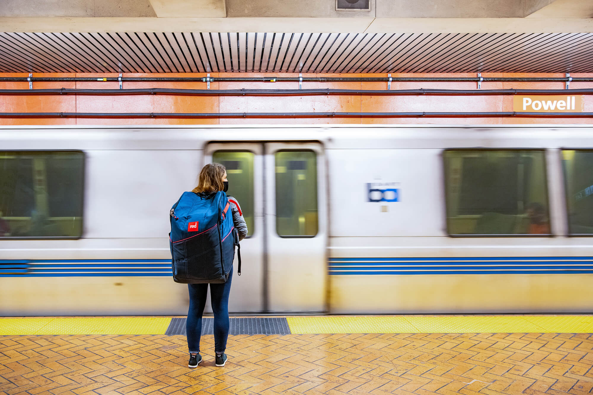 Woman With SUP Carrier On Her Back On A Powell Street Station Platform