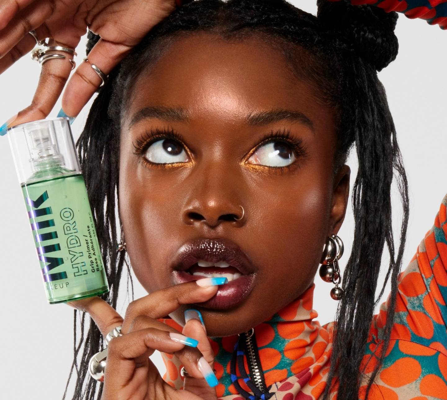 Model in a colorful outfit holds a bottle of Milk Makeup Hydro Grip Primer near her face, against a white background