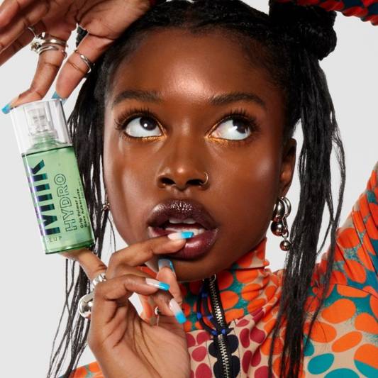 Model in a colorful outfit holds a bottle of Milk Makeup Hydro Grip Primer near her face, against a white background