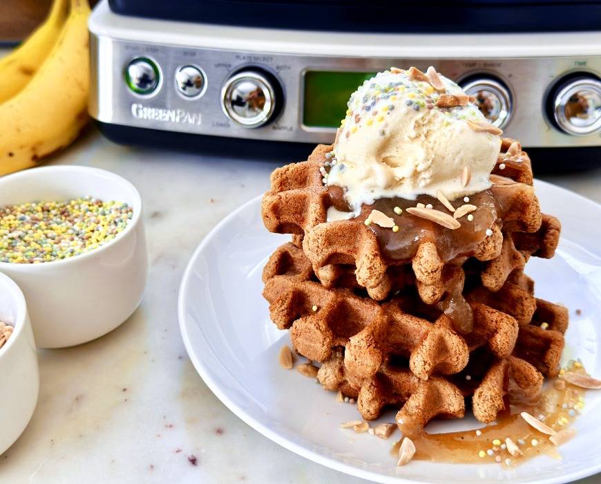 Stack of waffles topped with ice cream in the foreground, with bowl of sprinkles, bananas, and GreenPan waffle maker in the background