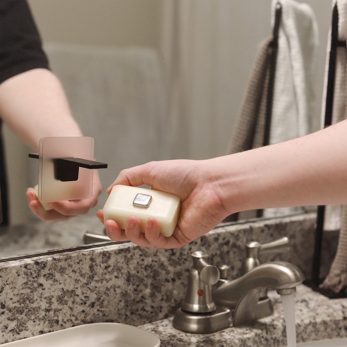 A hand holding a bar of soap next to the Traceless Adhesive Magnetic Soap Holder by Yamazaki Home in black.