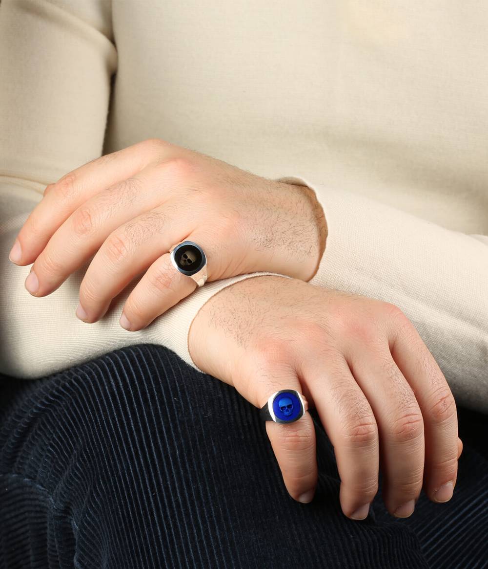 A man  is wearing two silver signet rings with black and blue enamel with a skull in the middle creating a ghost effect.