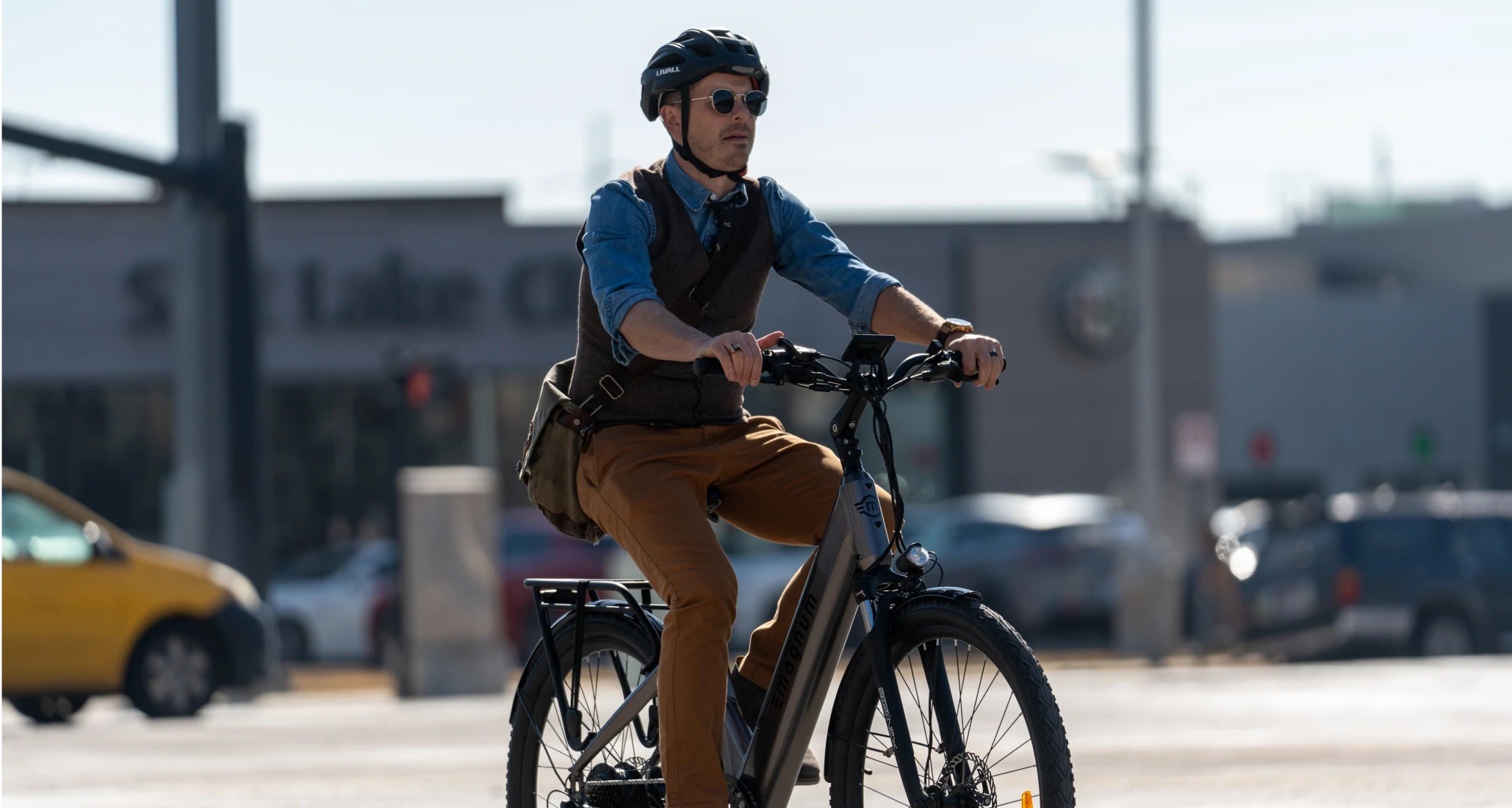 Man dressed in khaki pants brown vest blue shirt with sleeves rolled up and wearing sunglasses and a black helmet, riding a Magnum Cosmo+ e-bike in the street