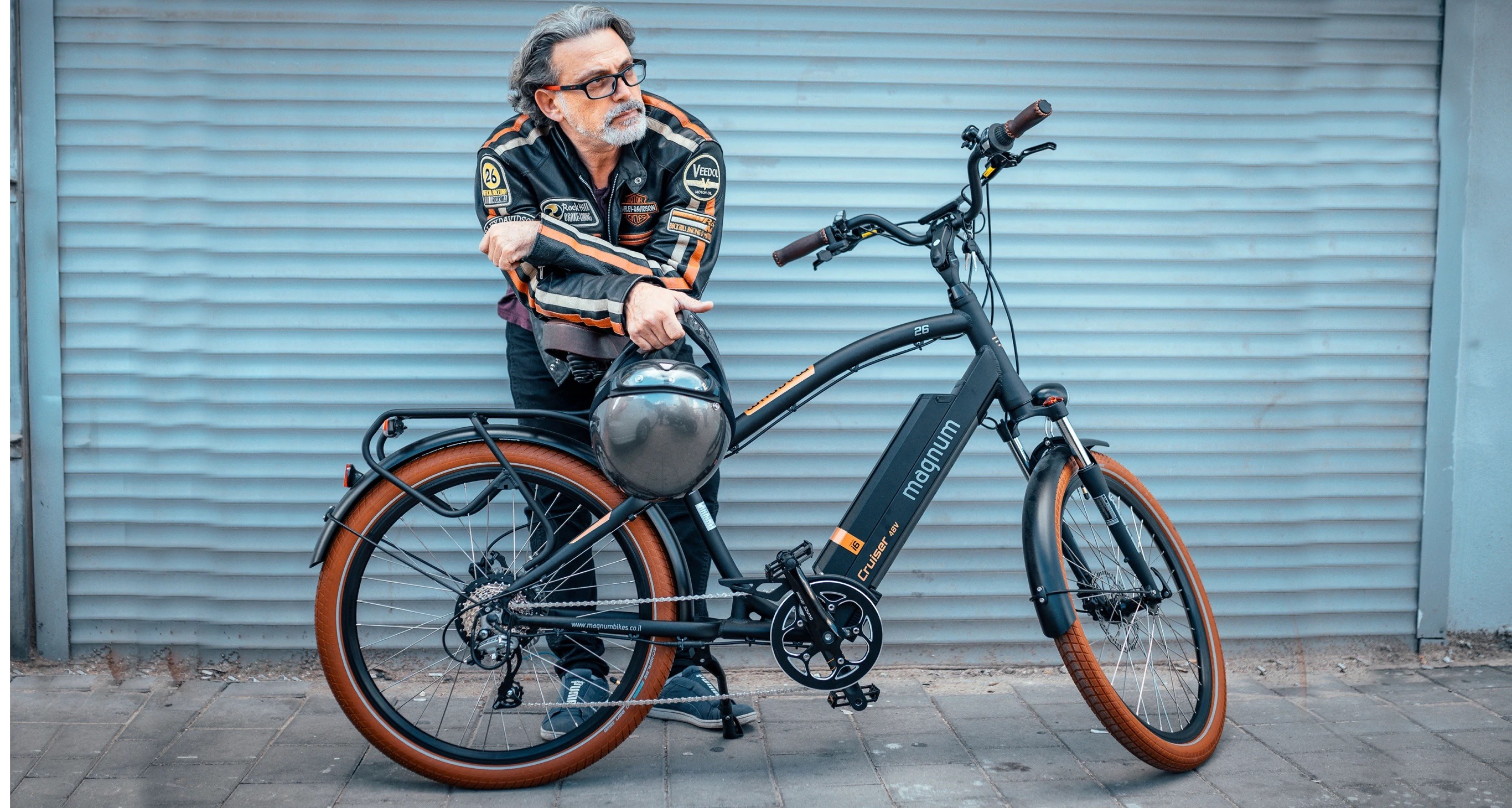 Man with gray hair and goatee wears a black and orange motorcycle jacket and leans over a Magnum Cruiser electric bike