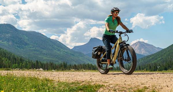 Woman in charcoal helmet turquoise shirt navy capris rides a Magnum Scout on a gravel trail in the mountains