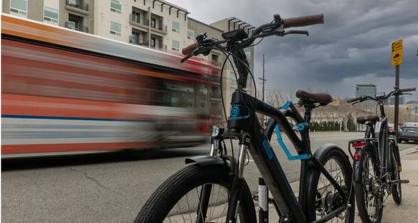 Black Magnum Voyager e-bike with blue accents and brown leather grips parked on a bike rack with a blurred bus whirring past in the background