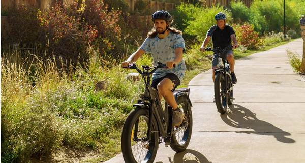 Two riders on Magnum Nomad electric bikes riding a paved path surrounded by plants