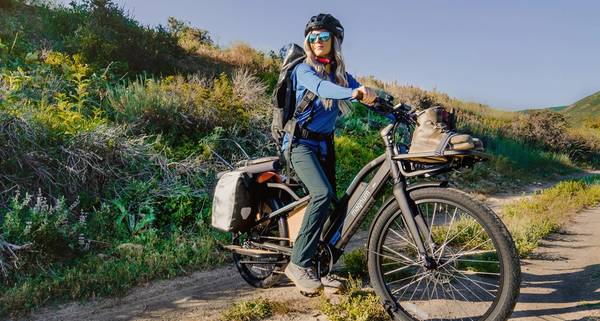 Rider with black helmet blonde hair blue reflective sunglasses blue jacket green pants on a dirt path straddling a Magnum Payload electric bike