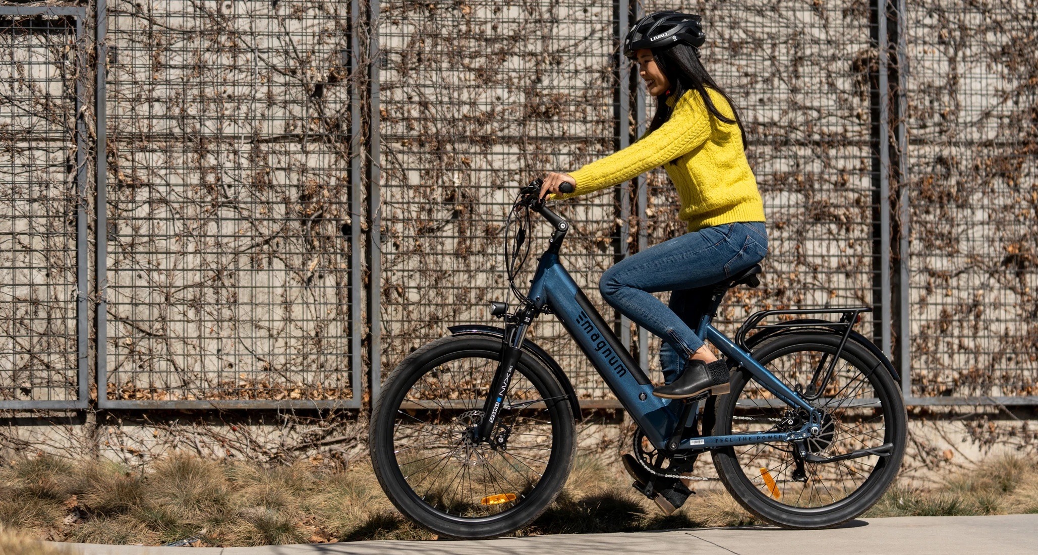Rider in yellow sweater and jeans on a blue Magnum Cosmo S riding in front of a black trellised wall