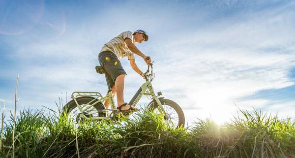 Rider in baseball hat striped polo shirt grey shorts and sandals riding Magnum Pathfinder 350 e-bike through the grass with sunny sky in background