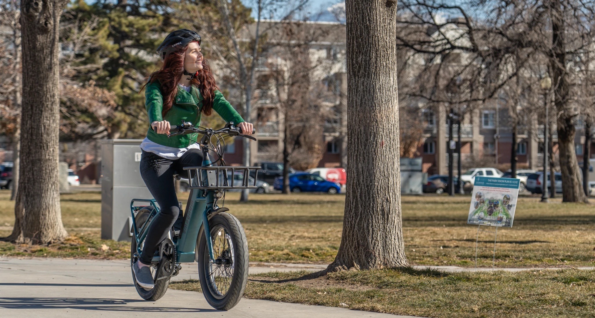 Rider with long auburn hair green jacket black pants smiles while riding a Magnum Pathfinder T electric bike through a park