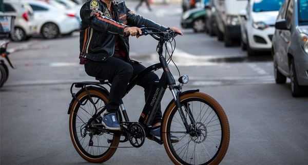 Rider in motorcycle jacket riding Magnum Cruiser e-bike across the street with parked cars in background