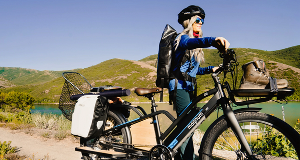 A woman with long hair steers a Magnum Payload cargo e-bike with a full load on front and back racks along a dirt path next to a lake