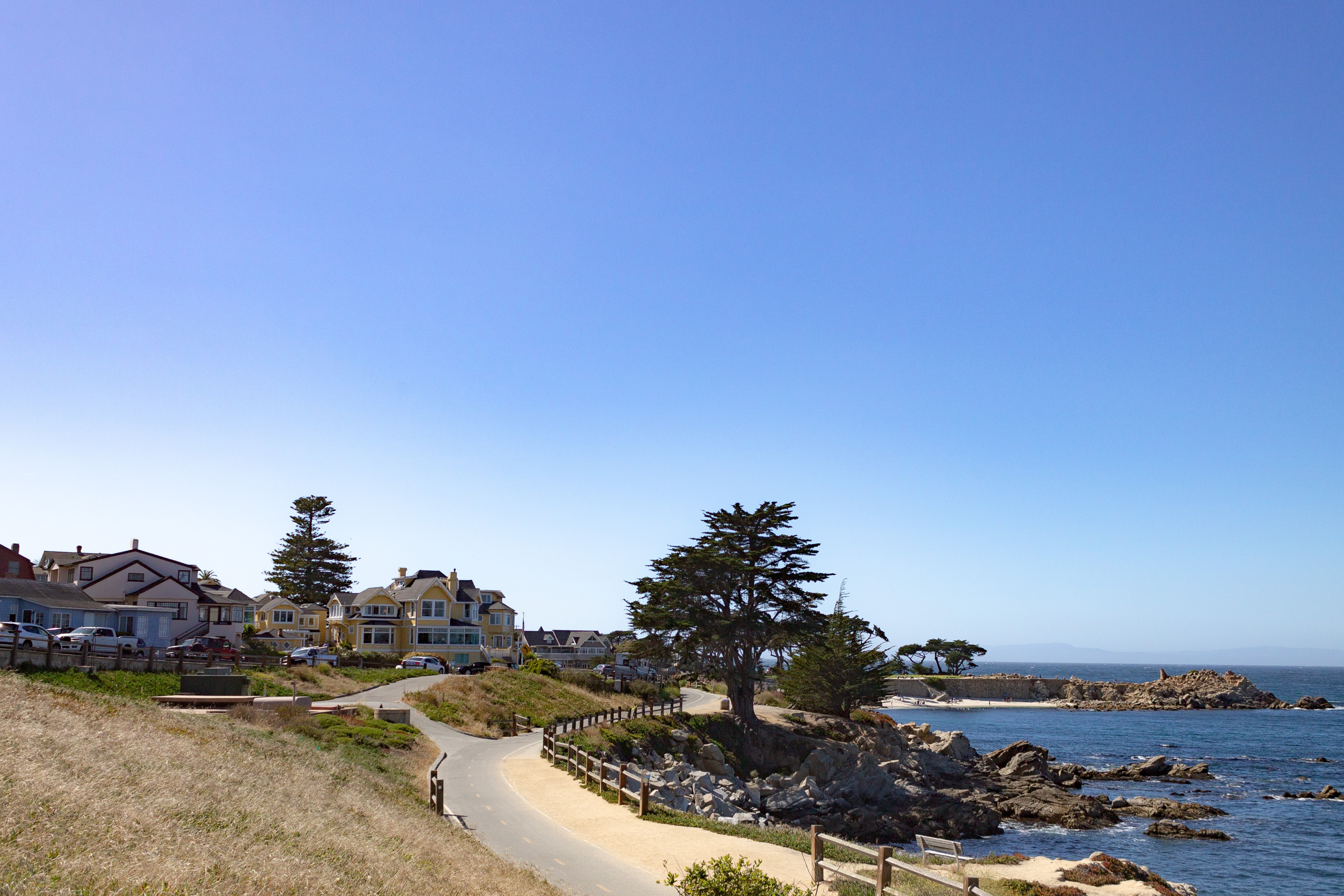 Clear blue sky with rocky ocean coastal view, a paved e-bike trail, dry summer grass, and buildings in the background
