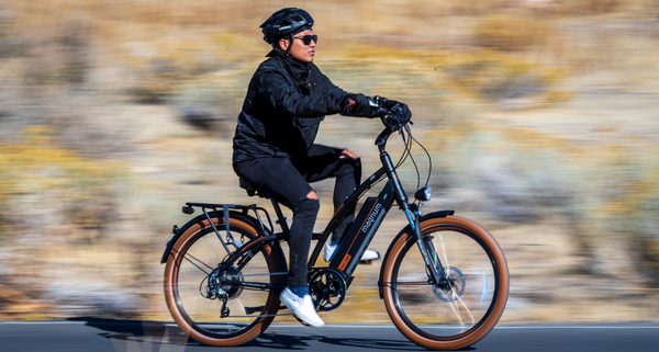 An e-bike rider dressed in all black with black jeans ripped at the knee; the rider and Magnum Low Rider cruiser-style e-bike are in focus while the background is blurred to show movement