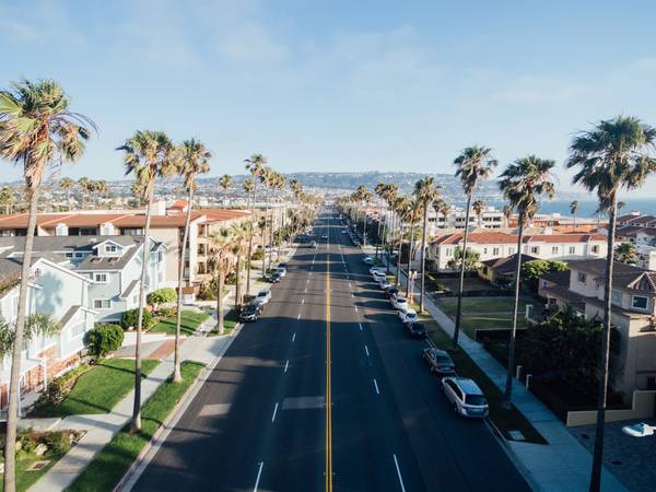 Four-lane road with cars parked on either side. Palm trees and buildings line the street and continue toward hills in the distance and a glimpse of ocean in the top right corner