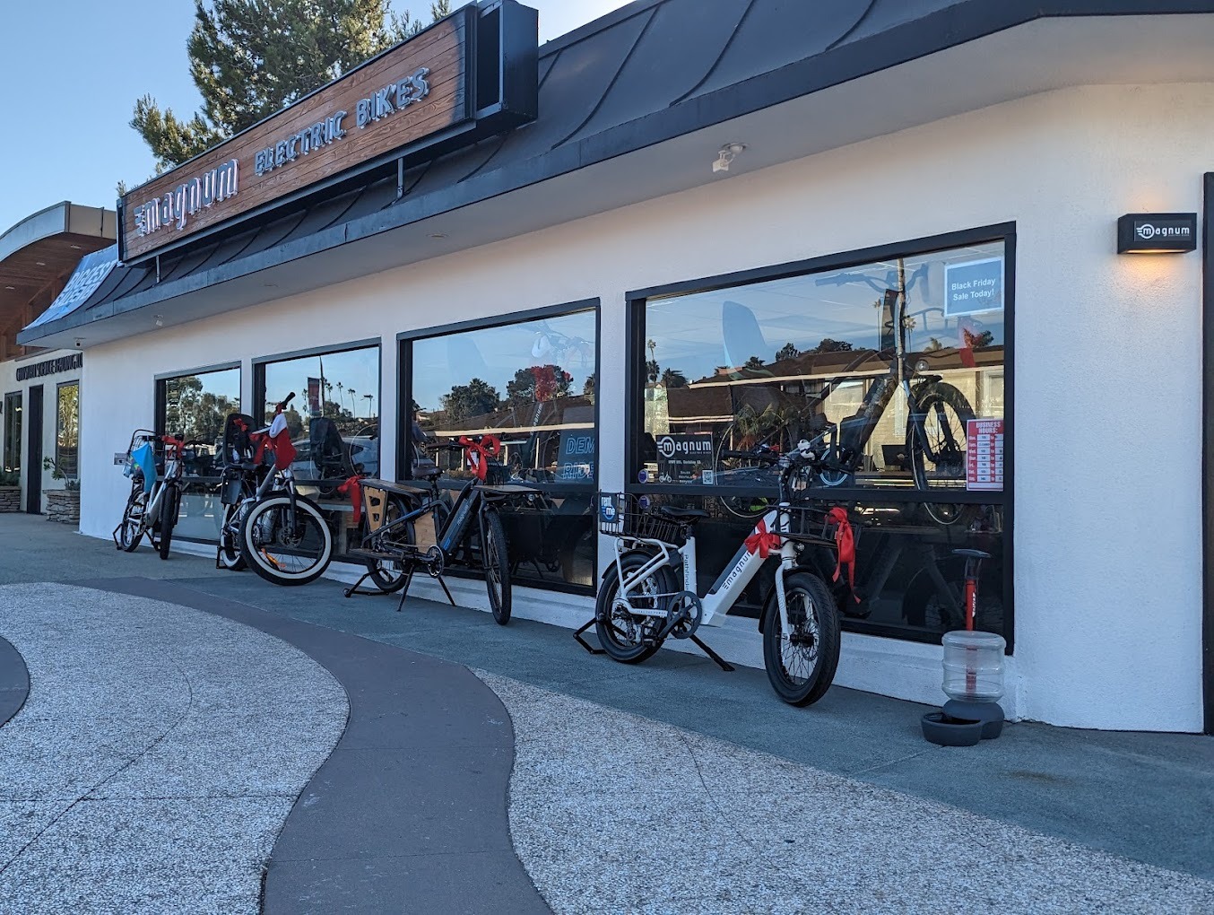 A selection of e-bikes posed in front of the Magnum Bikes flagship store location in Encinitas CA, featuring a cream-colored building exterior with black window frames and a wood background on the electric sign