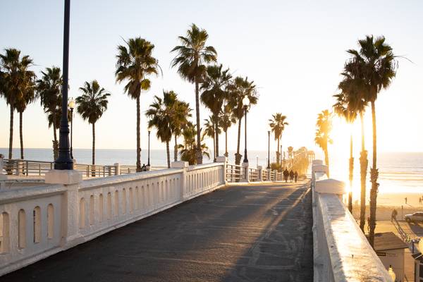 A concrete pier leading out to sunset over the ocean; palm trees, sand, and people are seen in the distance