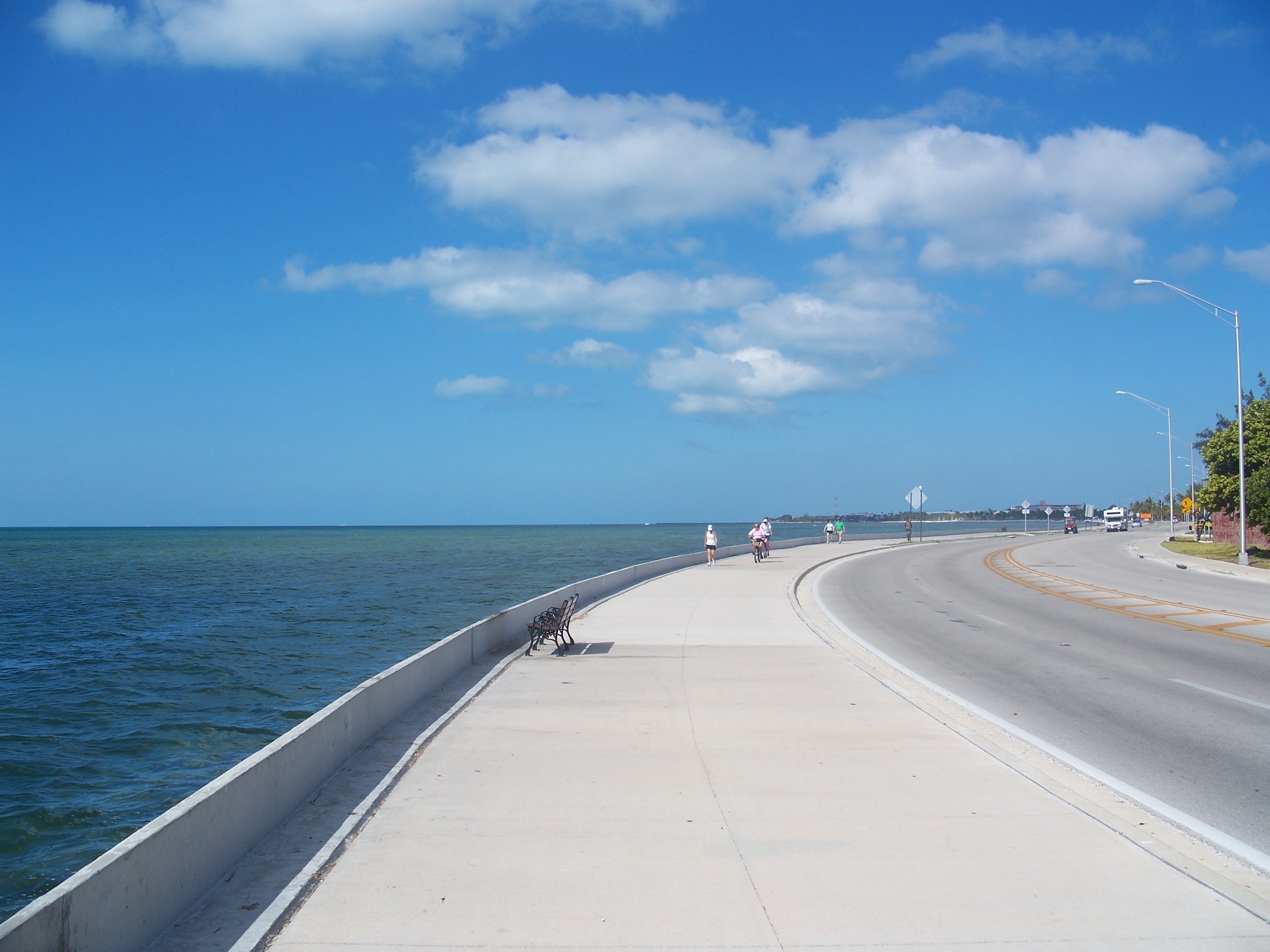A wide paved path and road curving around the coastline where blue water meets blue sky, with e-bikes and pedestrians in the distance