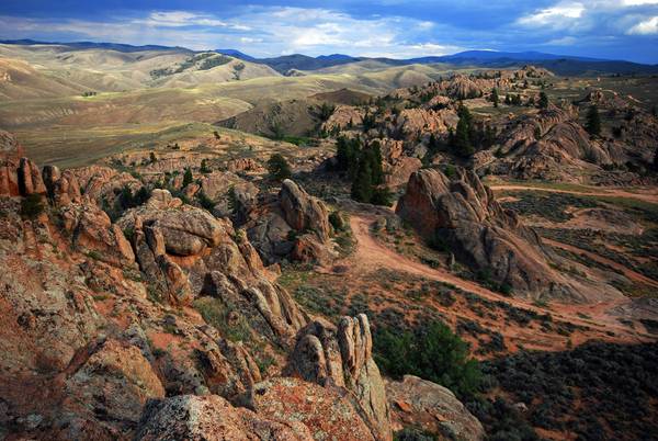 Red rocky Colorado landscape with trails, scattered greenery, green hills, and cloudy skies in the distance