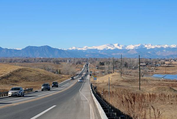 A long stretch of road in Colorado with brown hills on either side, tall hills in the distance, and snowy mountains in the background