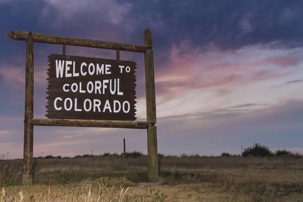 A purple, pink, and yellow cloudy sky over a dry field with a large wooden sign reading "Welcome to Colorful Colorado"